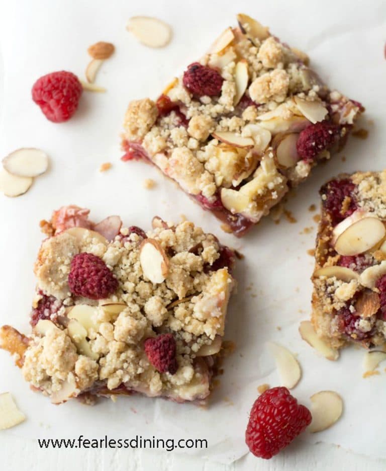 Three raspberry bars on a plate with fresh raspberries and sliced almonds.