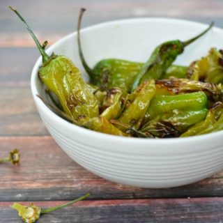 A large white bowl of pan fried shishito peppers on a wooden table.