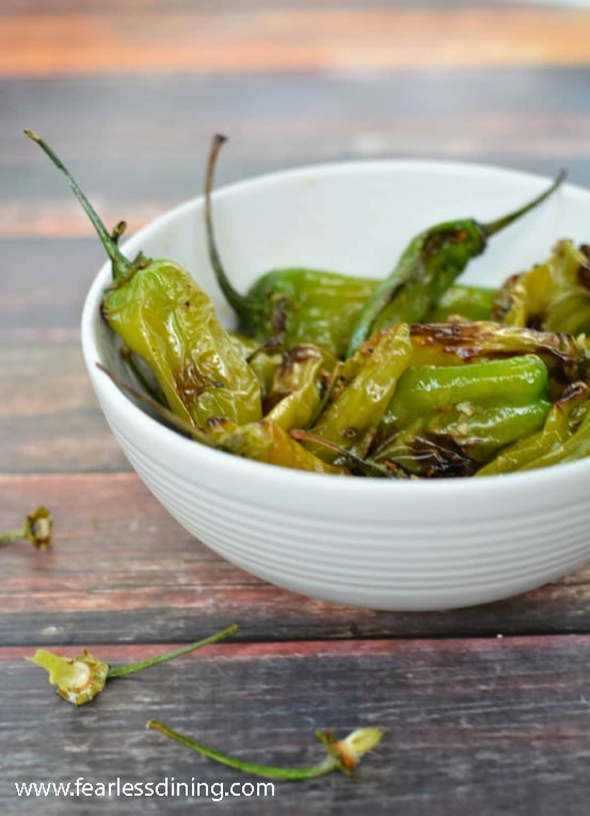 A bowl of pan fried shishito peppers on a wooden table.