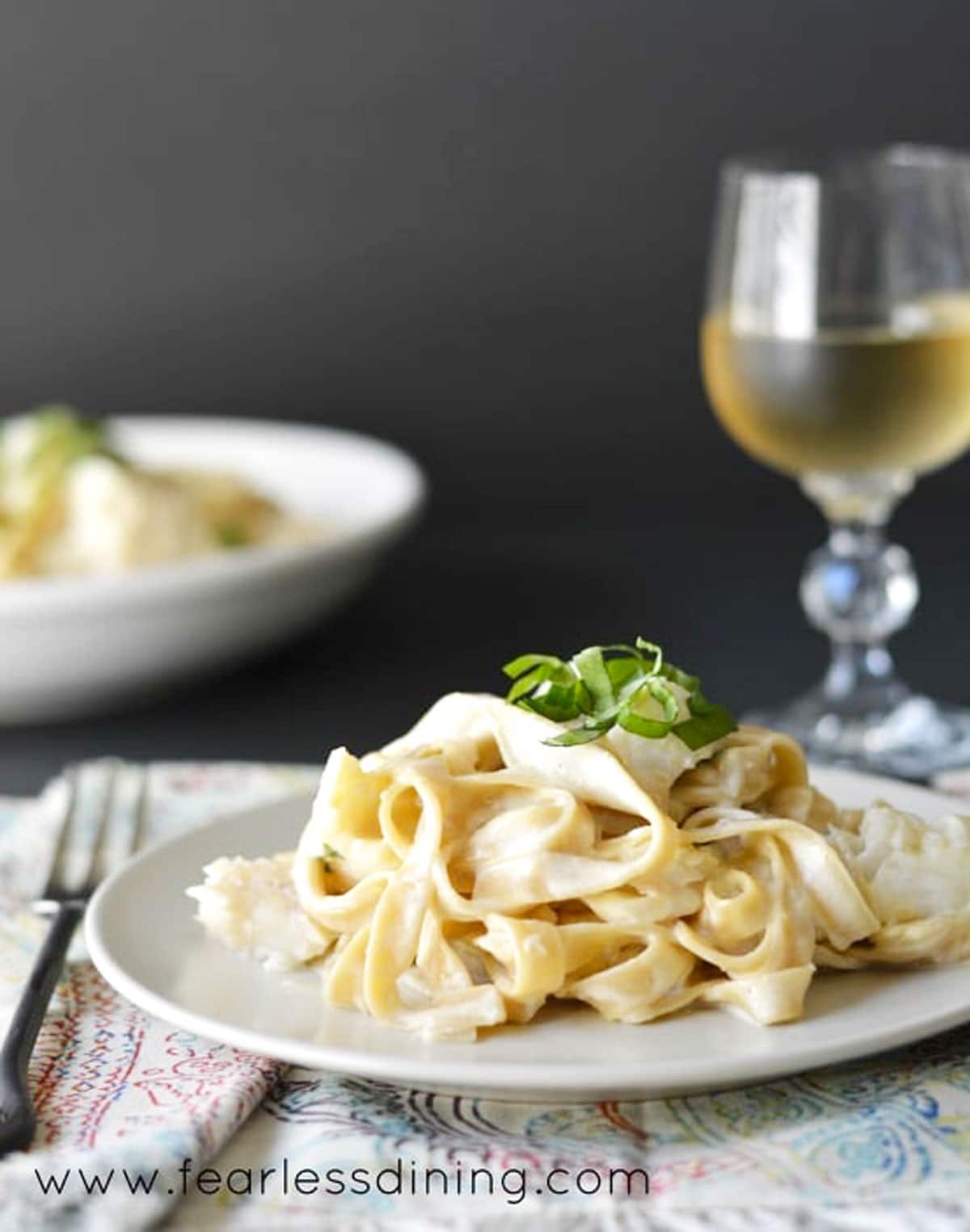 A plate full of cod fettuccine alfredo next to a glass of white wine.