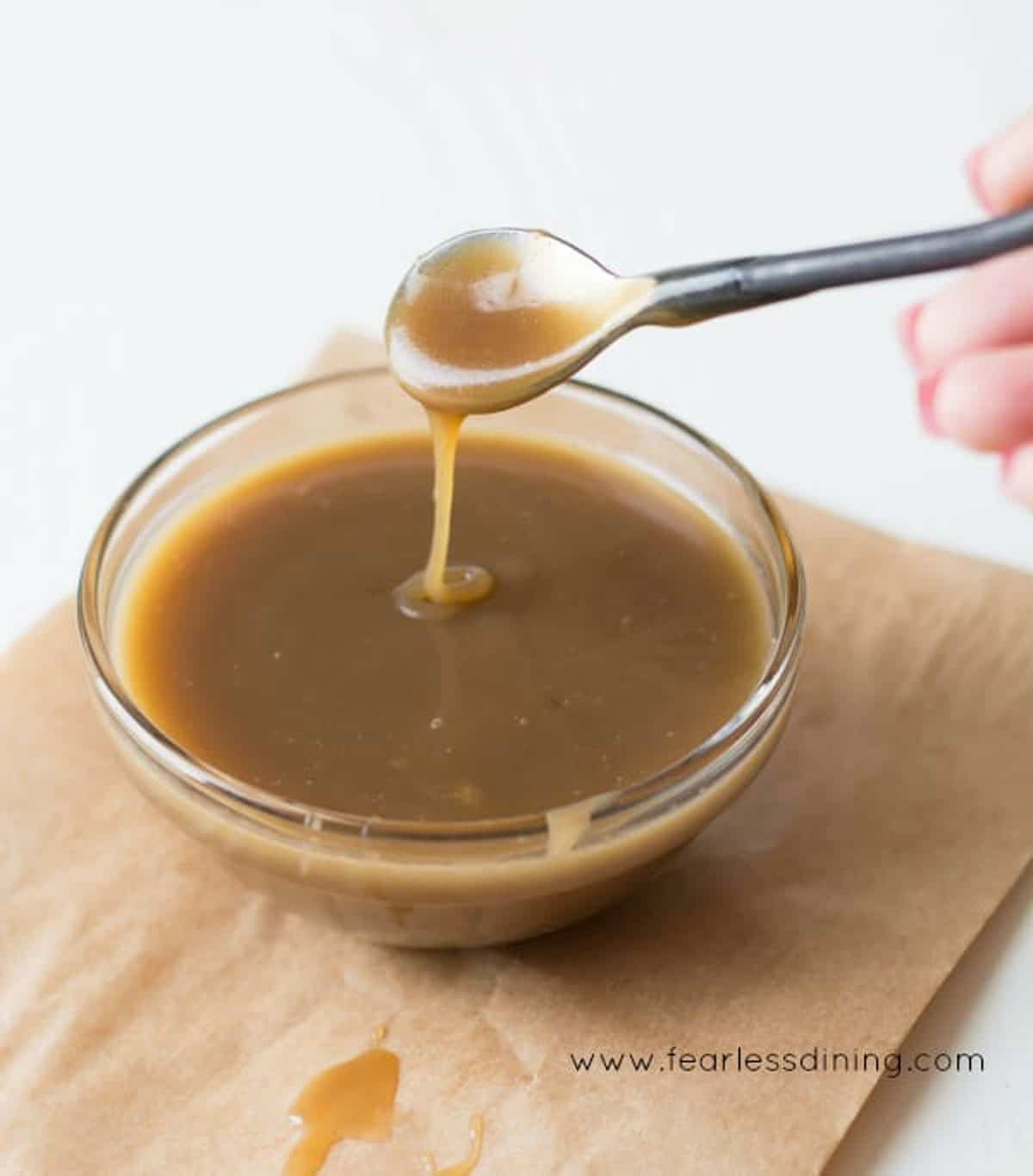 A glass bowl filled with butterscotch sauce. A spoon is drizzle the sauce into the bowl.