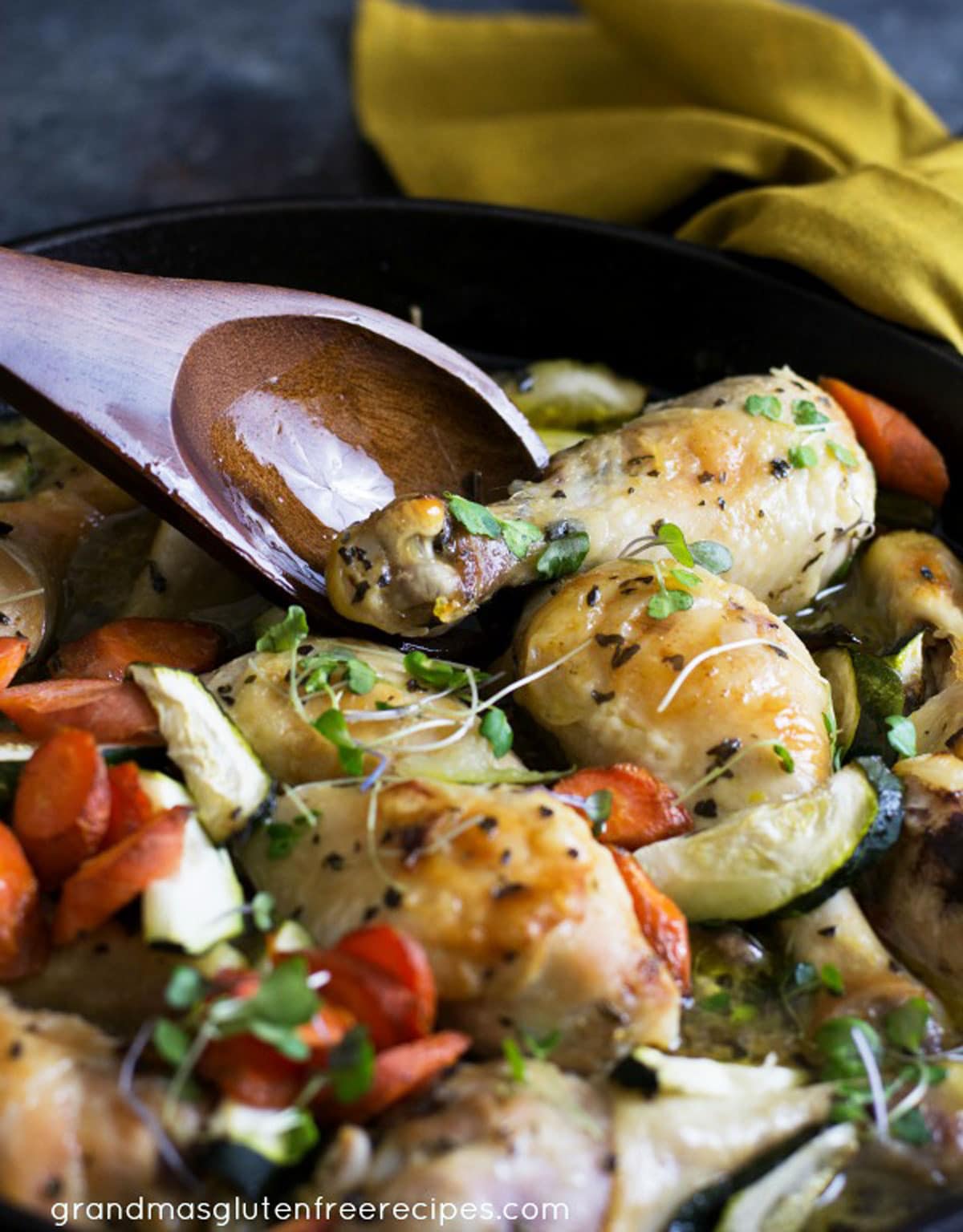 A wooden spoon lifting a chicken leg out of the skillet.