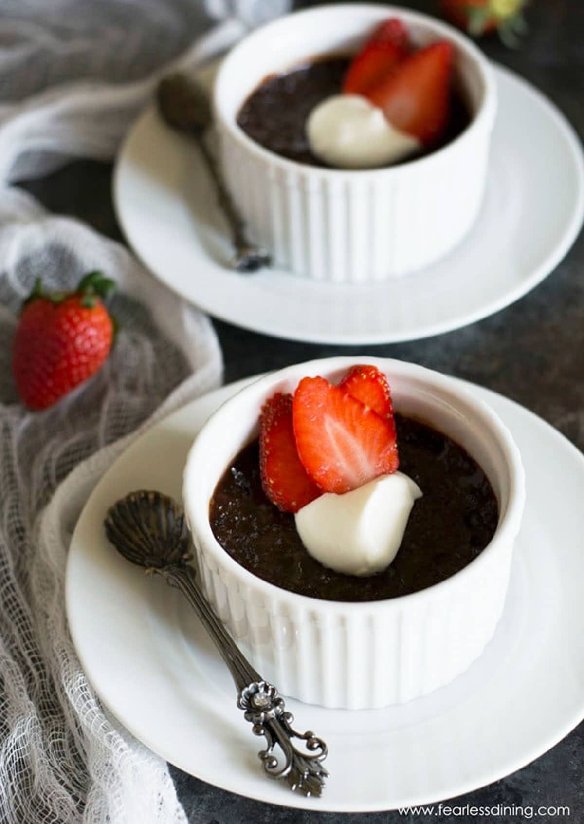 Two ramekins filled with chocolate custard. They are topped with whipped cream and sliced strawberries.