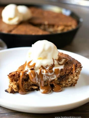 a slice of skillet cookie on a plate. It is topped with a scoop of ice cream and hot caramel.
