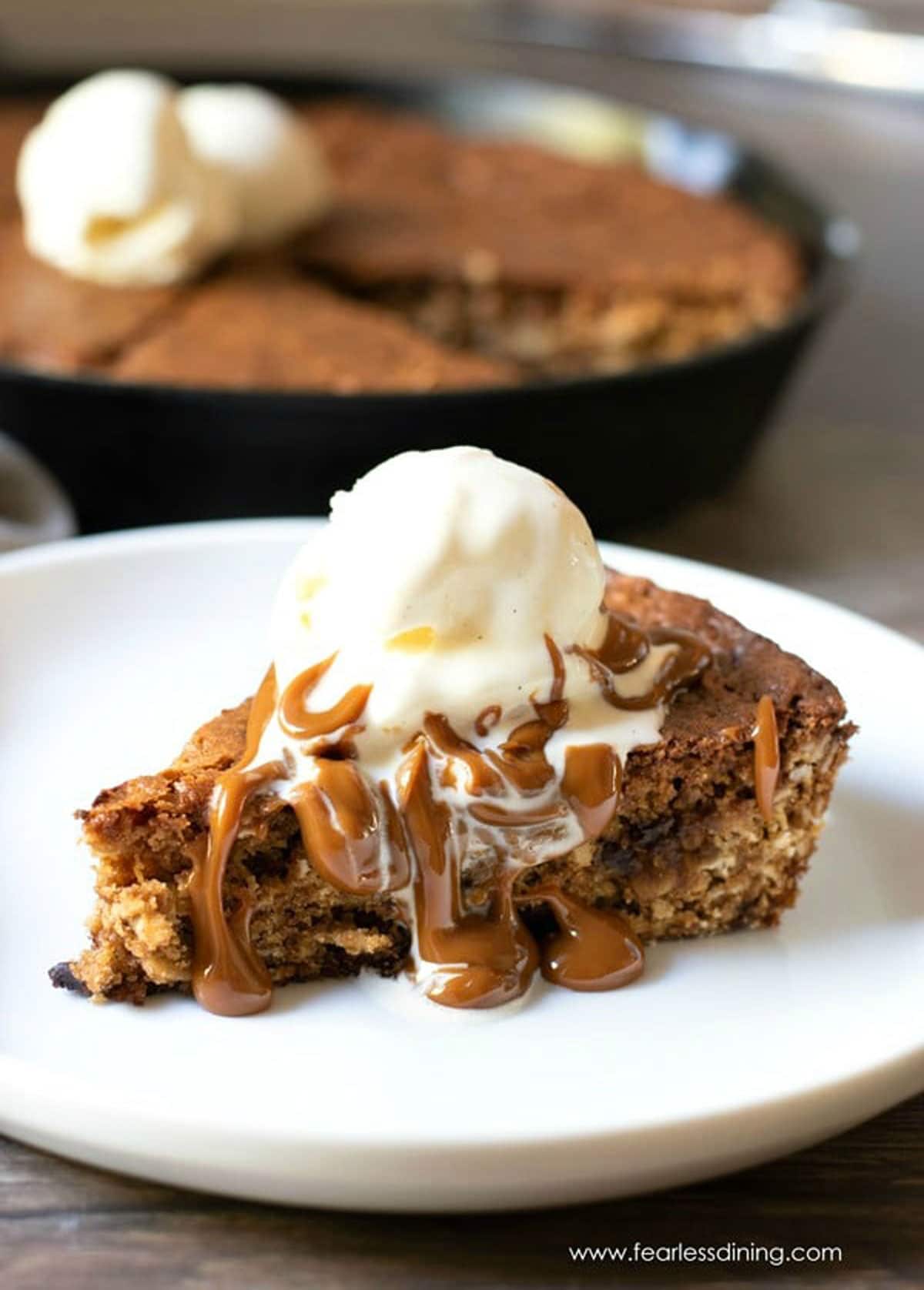 A slice of skillet cookie on a plate. It is topped with a scoop of ice cream and hot caramel.