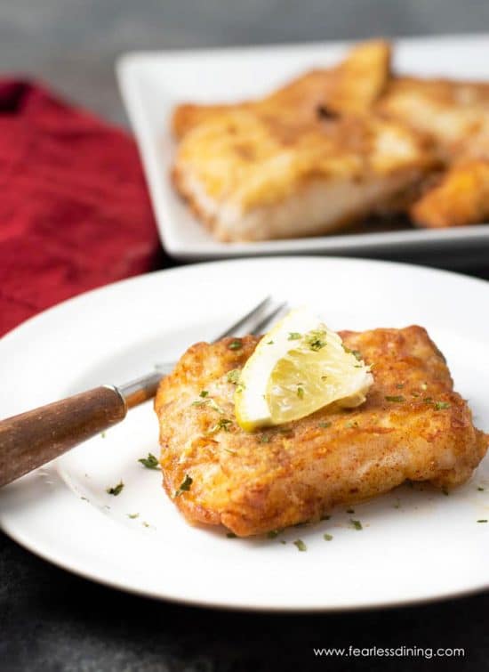 A white plate with a piece of air fried cod. The serving platter of cod is behind the plate.