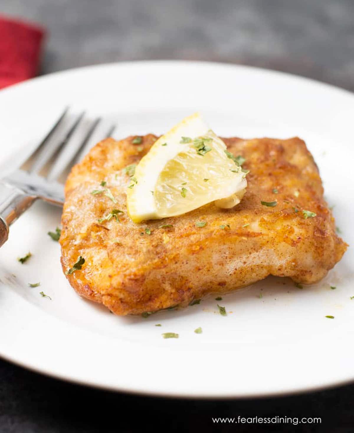 a single piece of air fried cod on a white plate. It is garnished with lemon and parsley