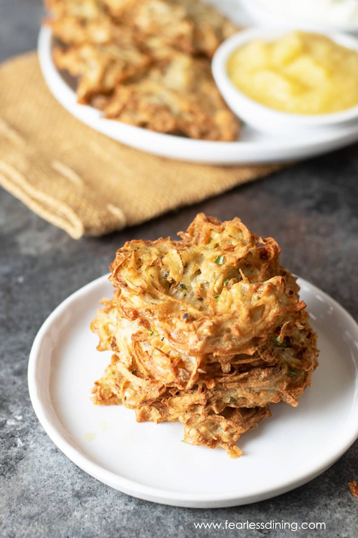 A stack of latkes on a white plate next to apple sauce.