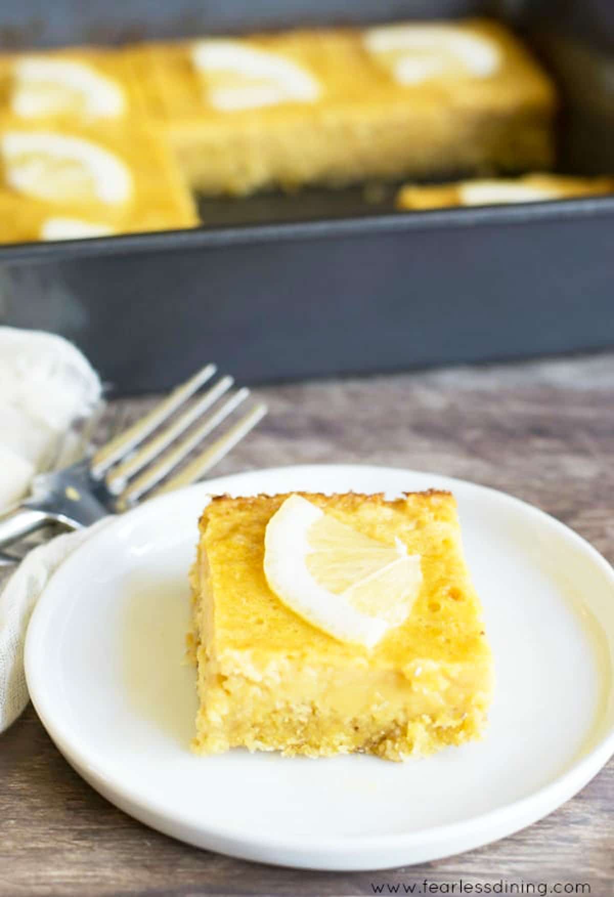 A lemon bar on a small white plate in front of the pan.