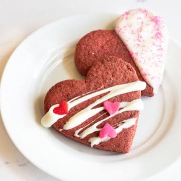 two decorated heart shaped red velvet cookies on a small white plate