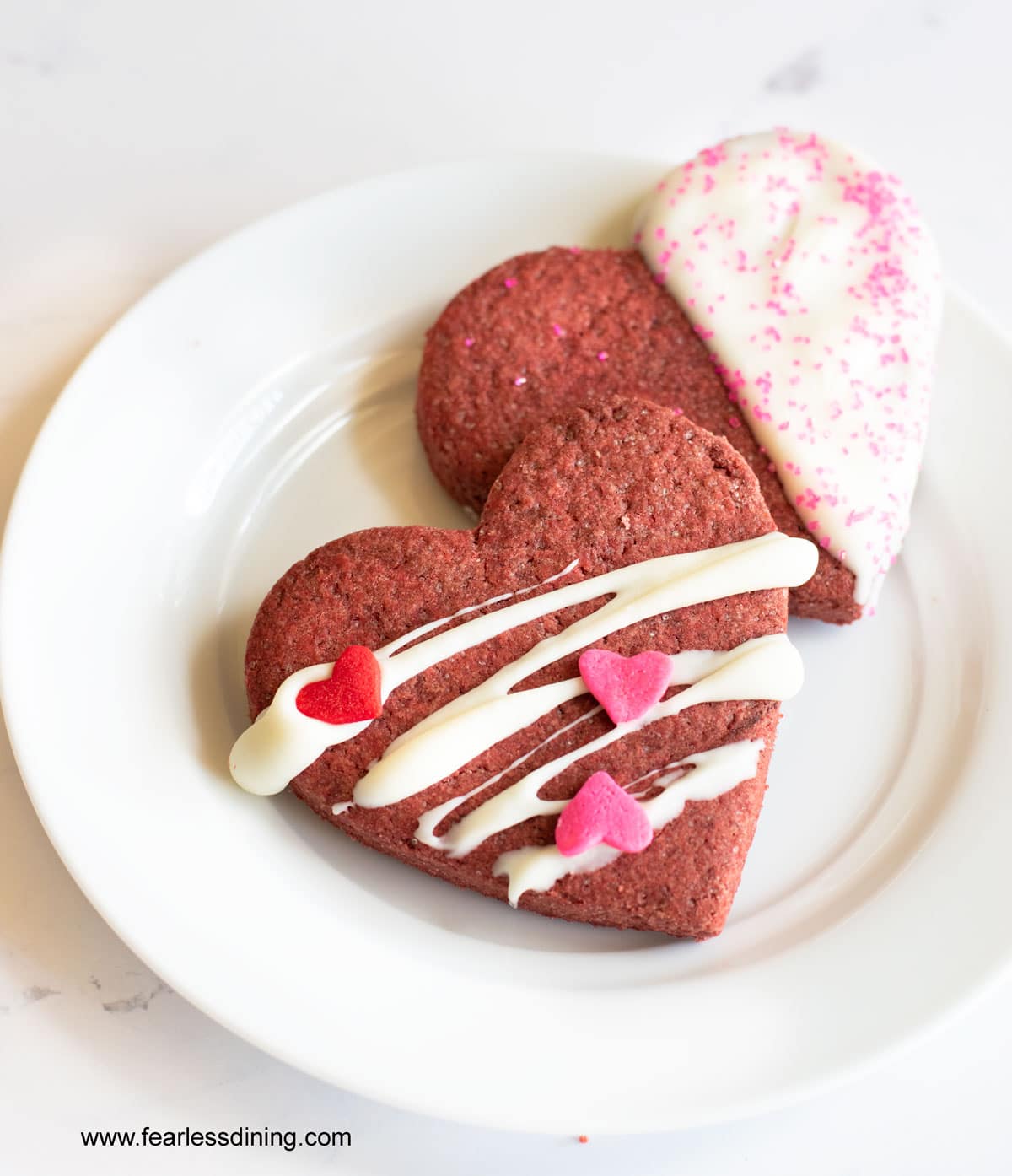 Two decorated heart shaped red velvet cookies on a small white plate.