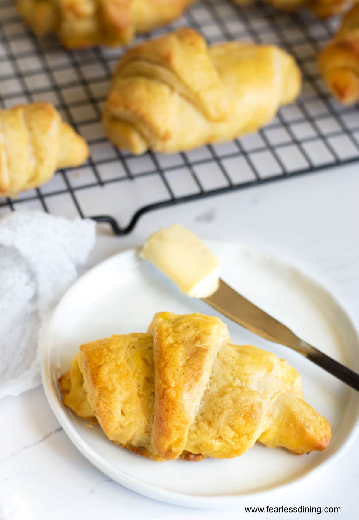 A gluten free crescent roll on a white plate next to a rack of crescent rolls.
