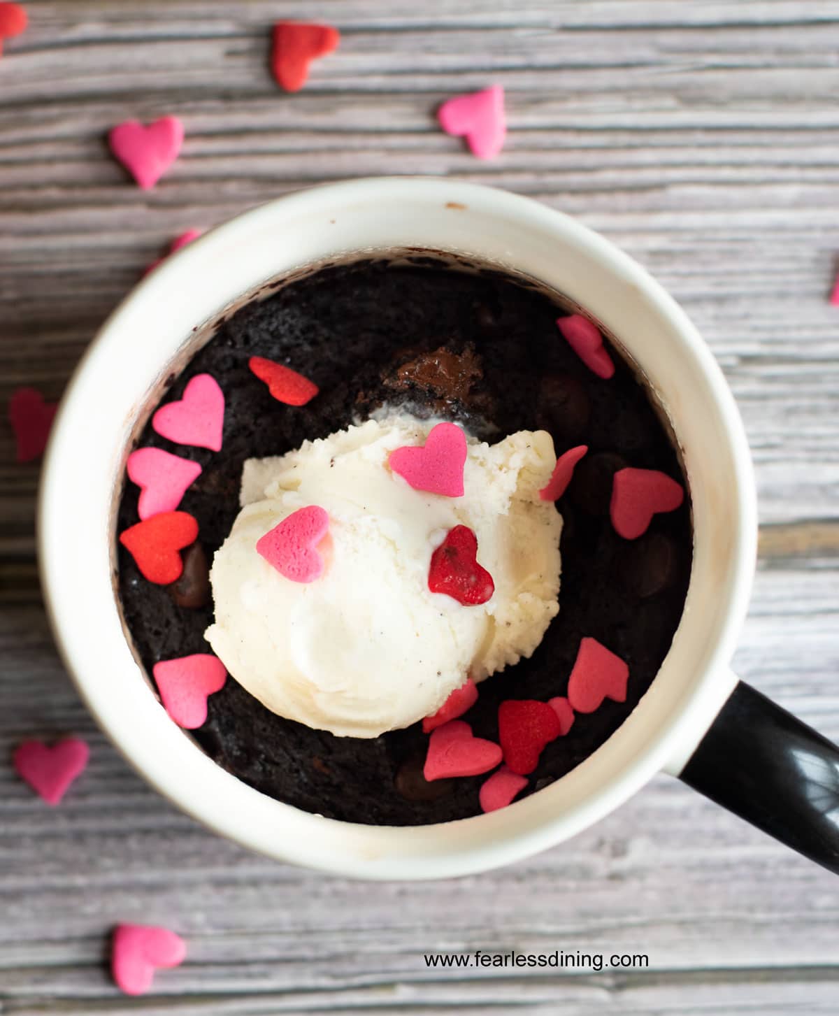 A cooked brownie mug with vanilla ice cream and pink and red heart sprinkles.