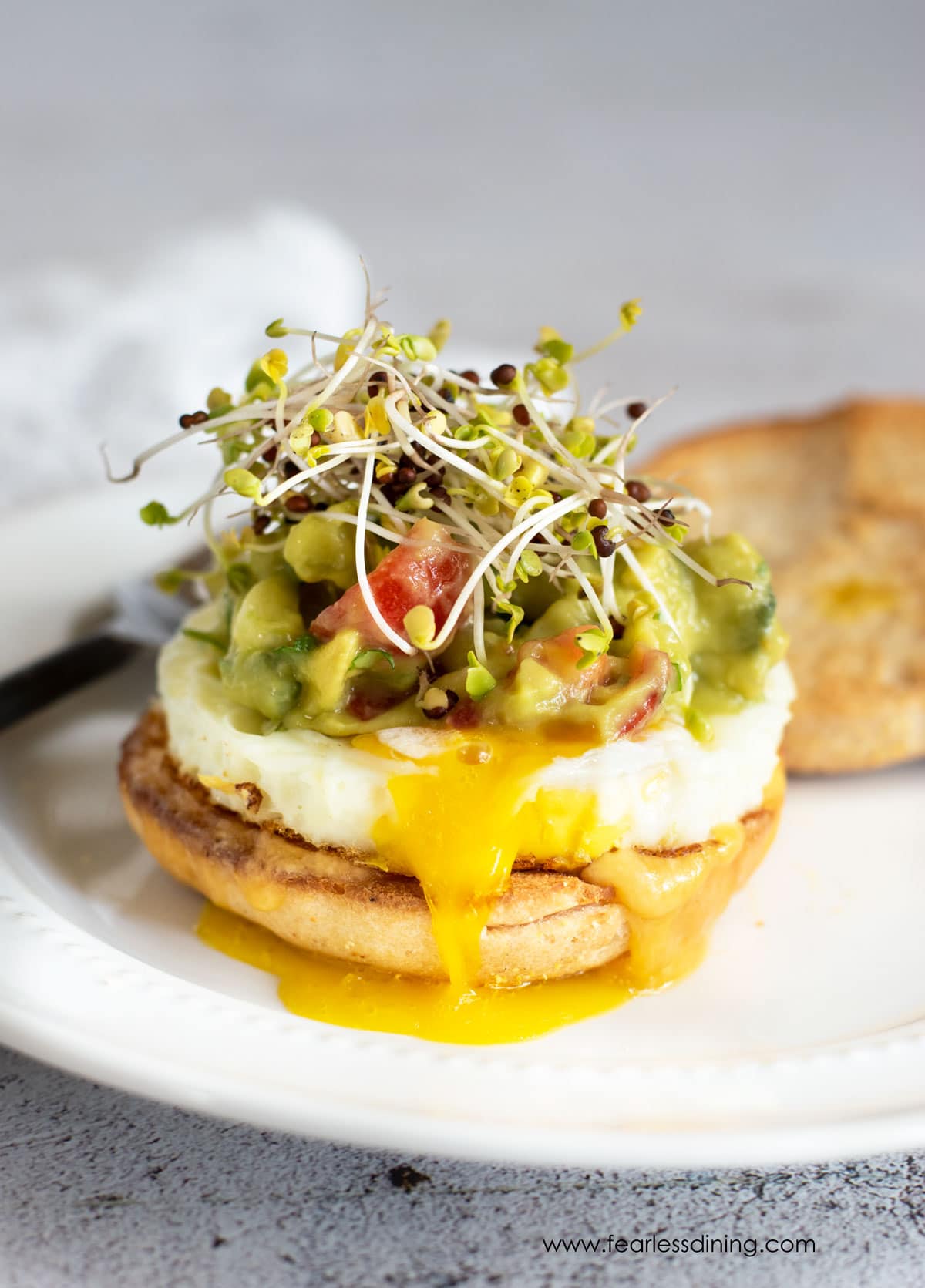 A gluten free breakfast sandwich topped with homemade guacamole and sprouts on a white plate.