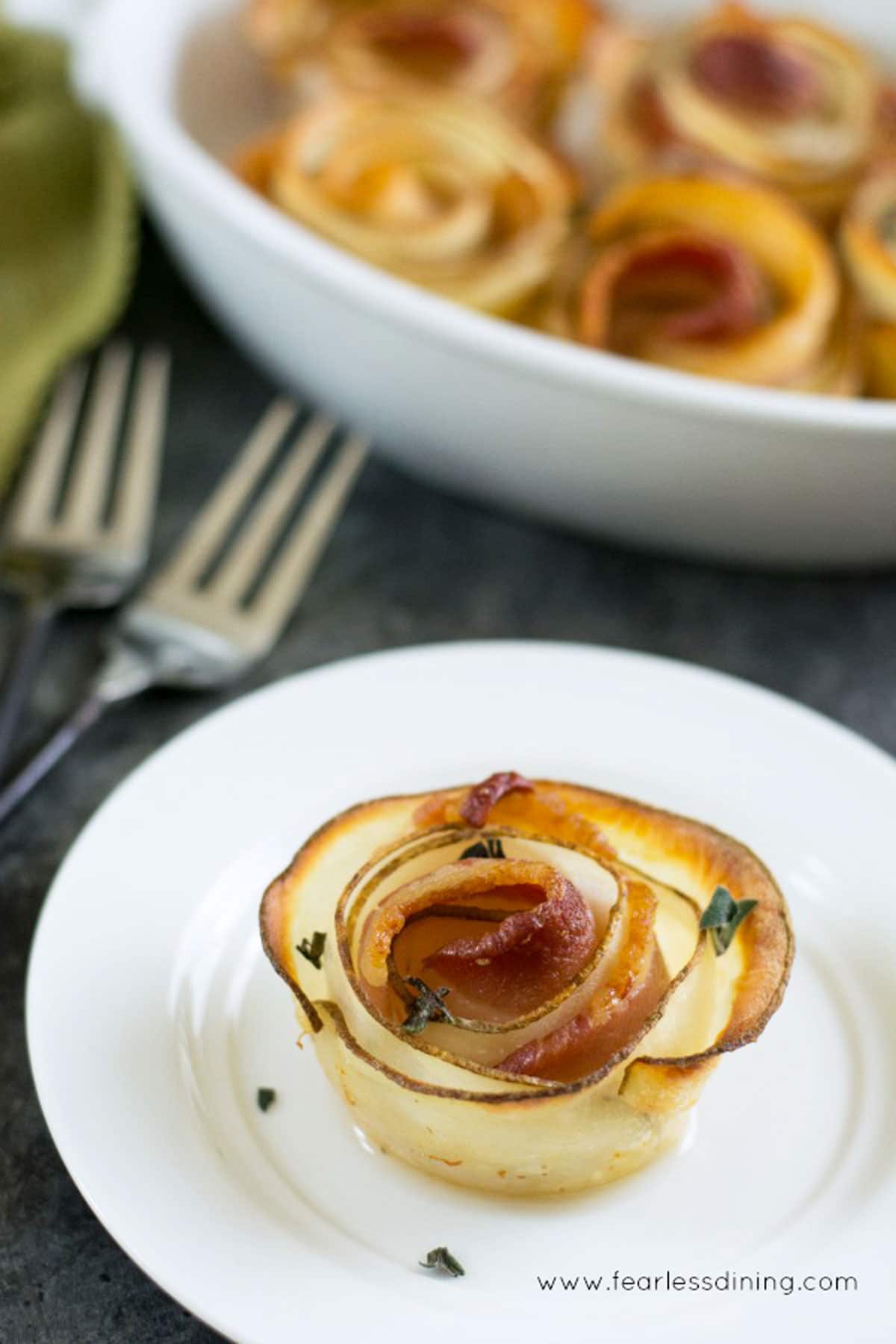 A bacon potato rose on a plate next to a baking dish full of bacon roses.