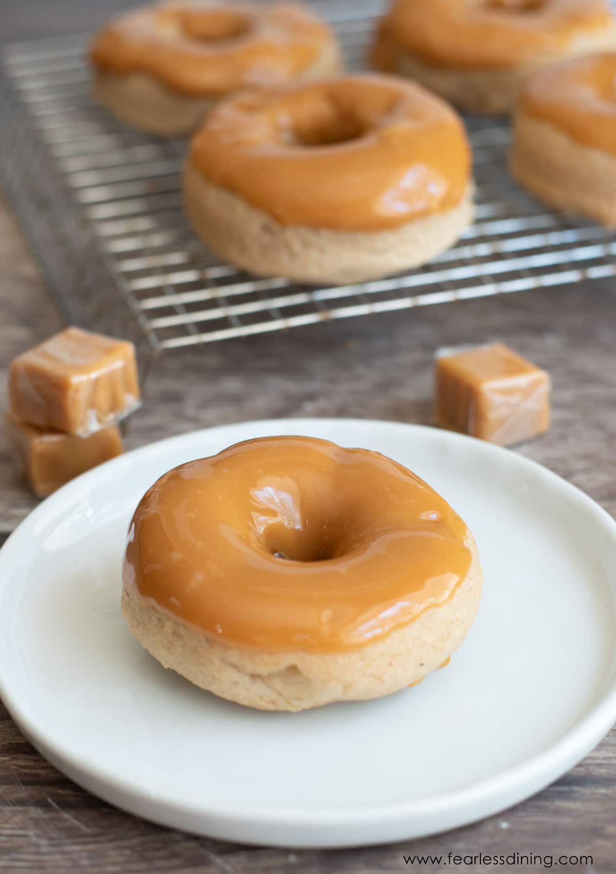 A caramel apple donut on a plate next to a rack of donuts.