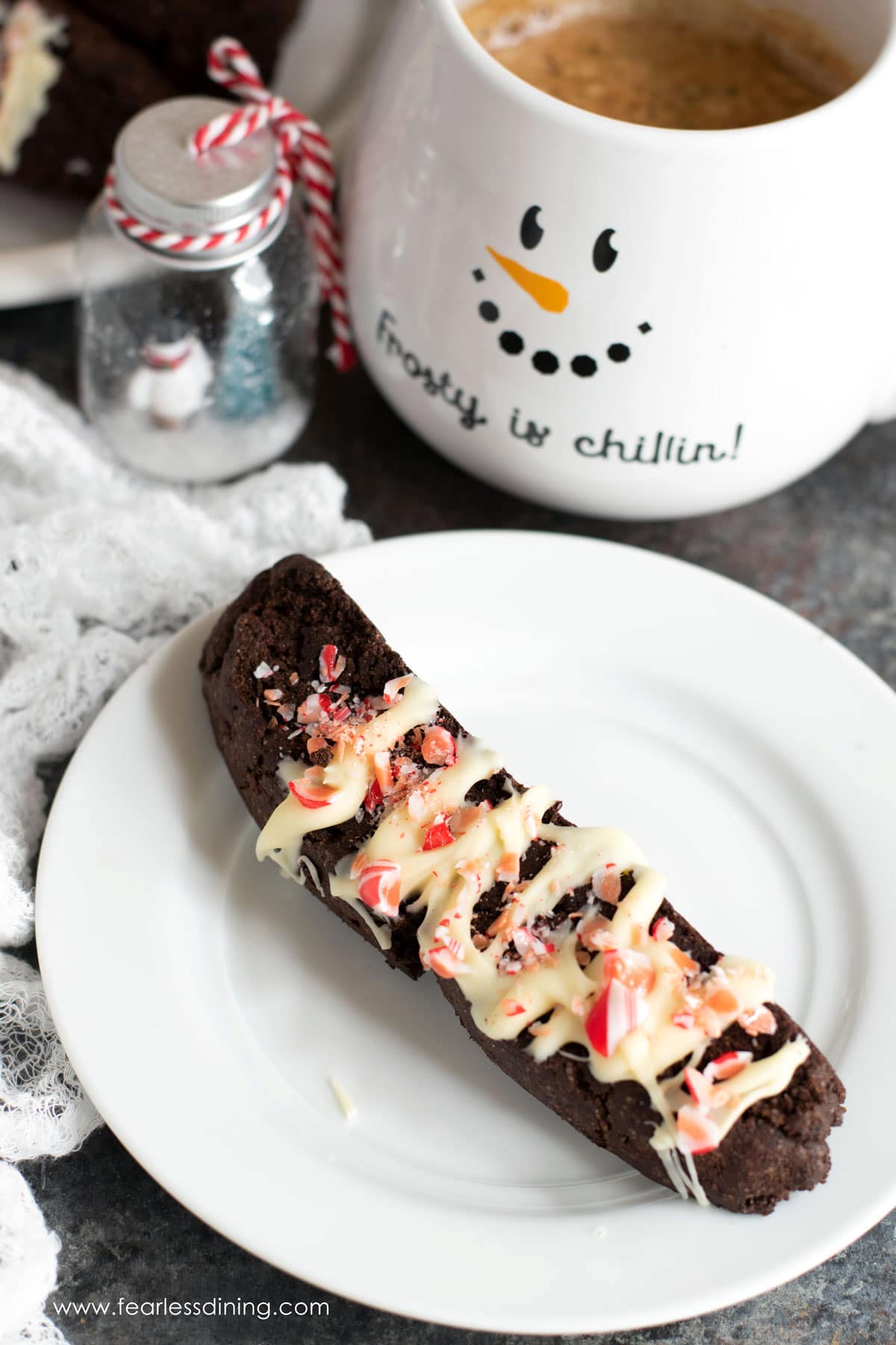 a piece of chocolate peppermint biscotti on a plate next to a snowman coffee mug.