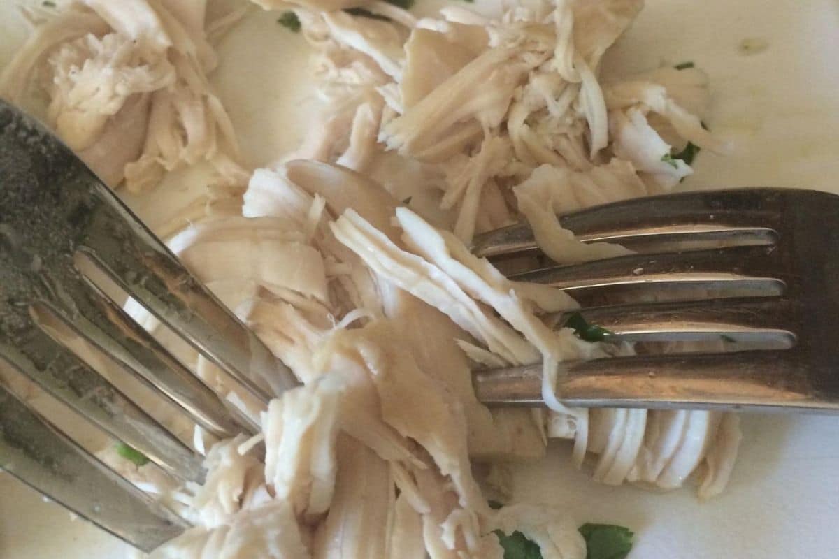 Shredding chicken breasts with two forks on a cutting board.