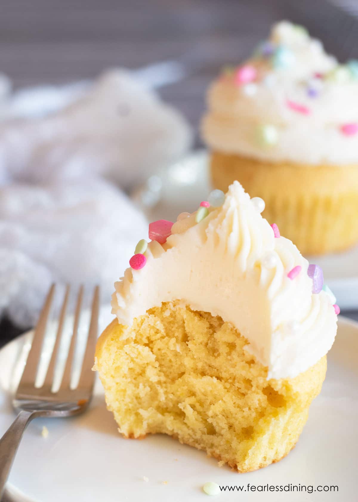 A frosted gluten free vanilla cupcake with a bite taken out so you can see the fluffy inside texture.