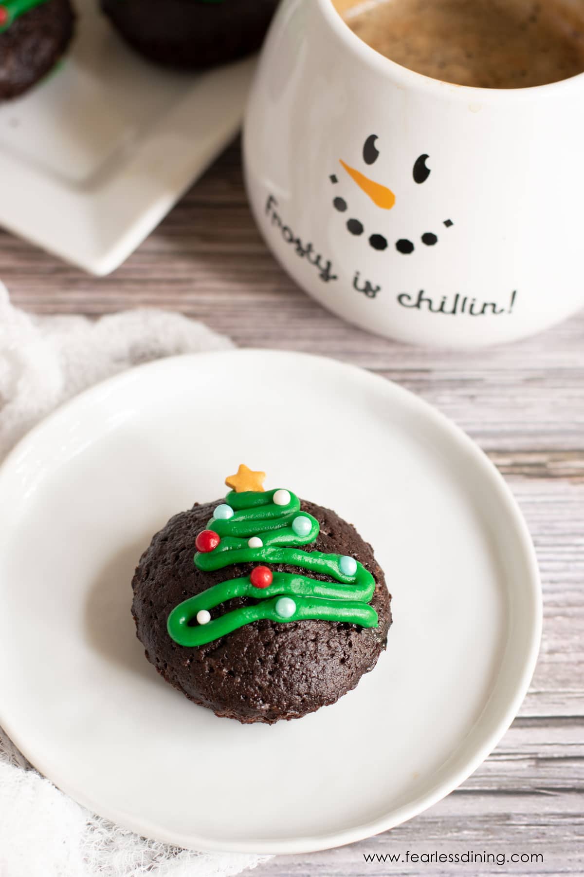 A Christmas tree decorated brownie bite on a white plate next to a cup of coffee.