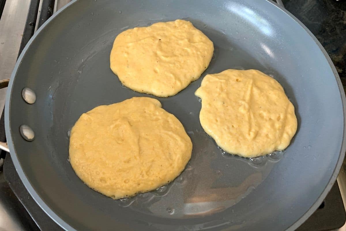 three cornmeal pancakes cooking in a frying pan.