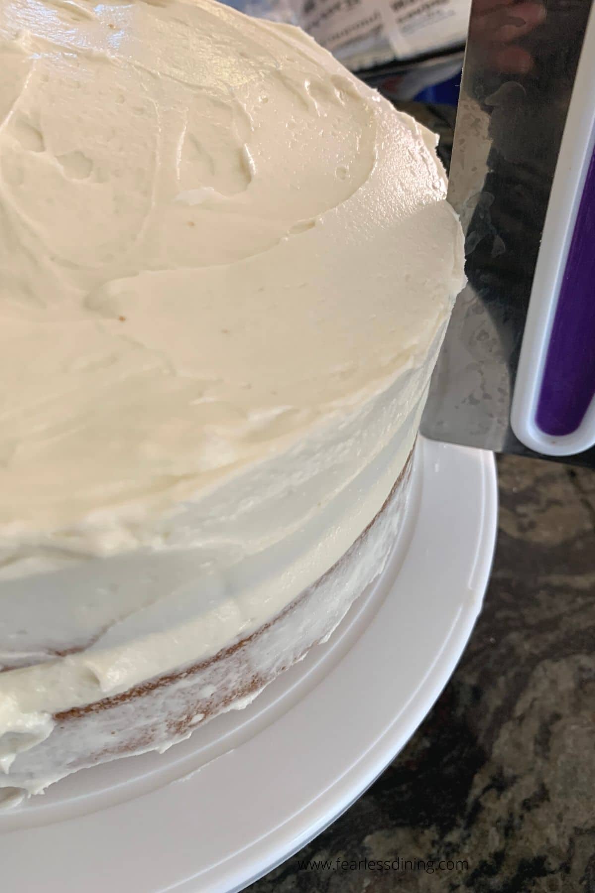 Using a scraper to scrape frosting from the side of the cake.