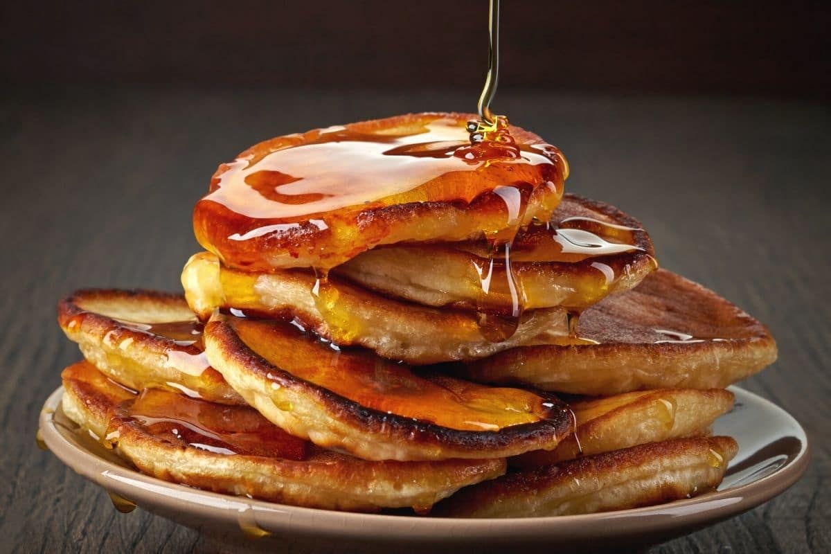 A stack of five pancakes with maple syrup being poured over them.