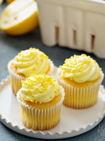 three lemon cupcakes on a plate. They are frosted and topped with lemon sprinkles.