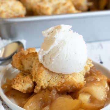 A serving of apple cobbler on a small white plate. It is topped with a scoop of vanilla ice cream.