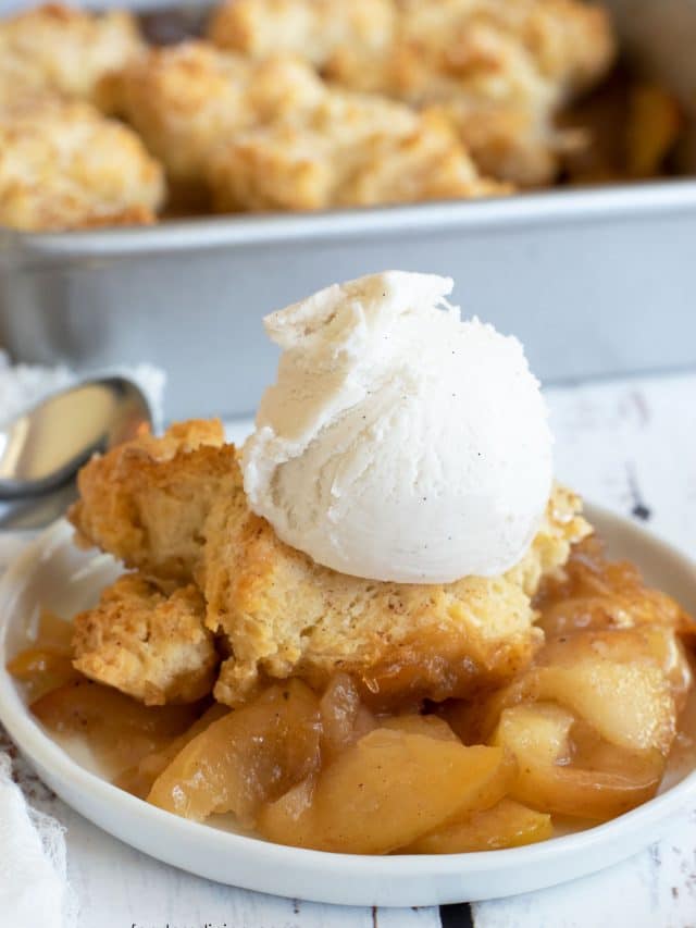 A serving of apple cobbler on a small white plate. It is topped with a scoop of vanilla ice cream.