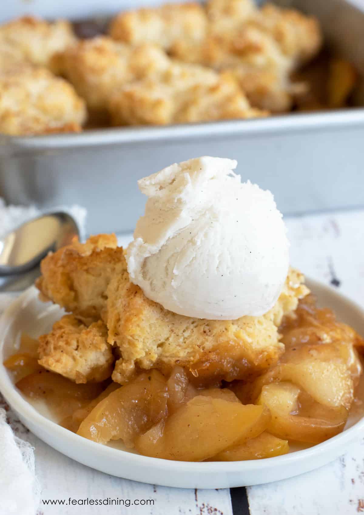 A serving of apple cobbler on a small white plate. It is topped with a scoop of vanilla ice cream.