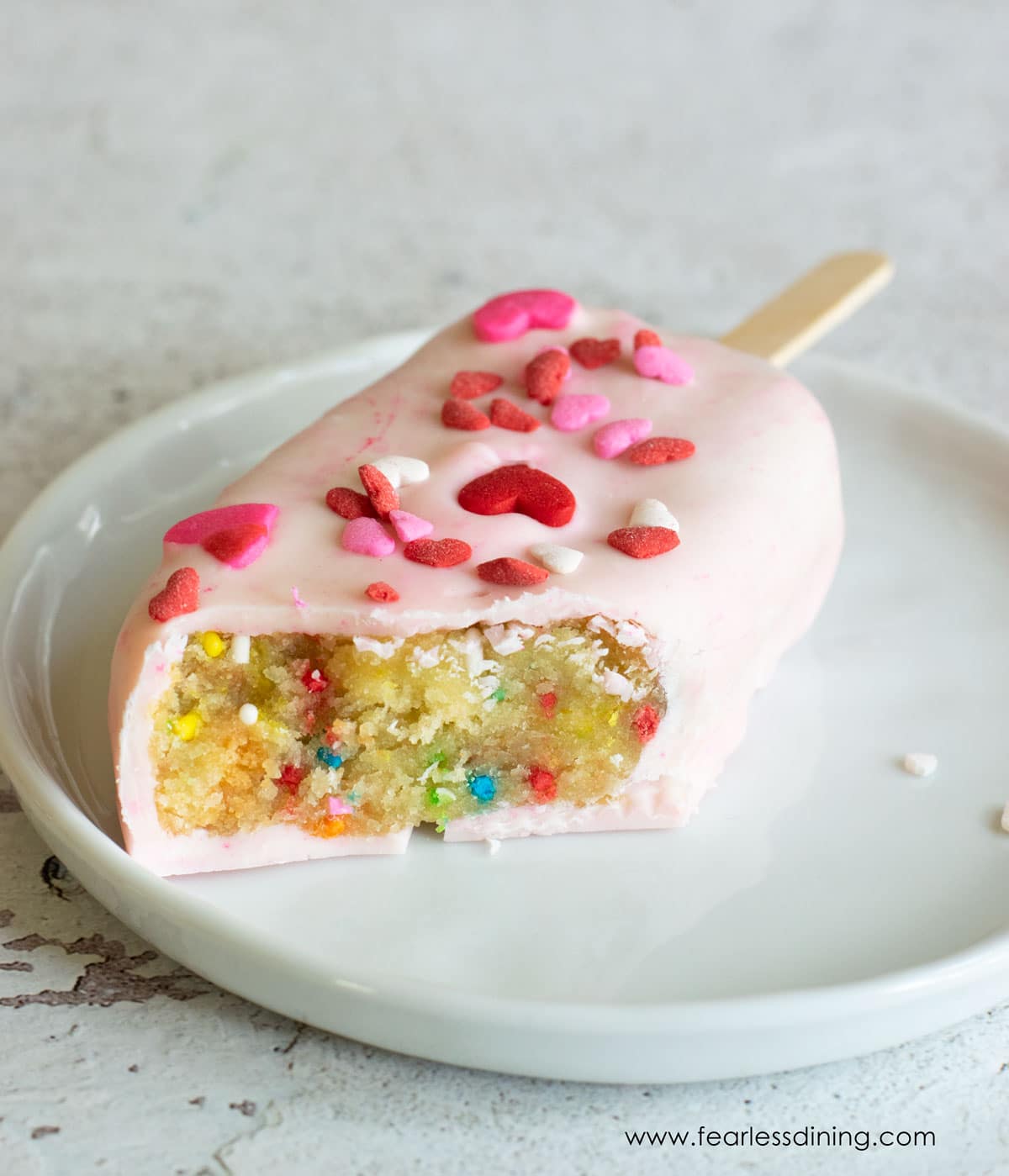 A pink candy coated funfetti cakesicle on a plate.