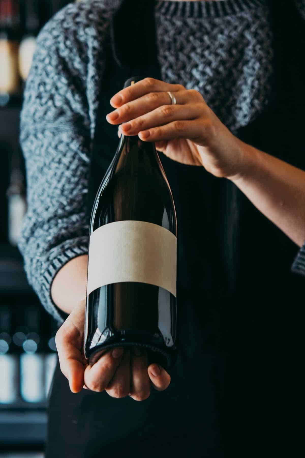 A person in a blue sweater holding up a wine bottle so you can see the label on the bottle.