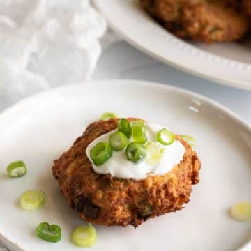 A salmon patty on a plate. It is topped with sour cream and fresh sliced scallions.