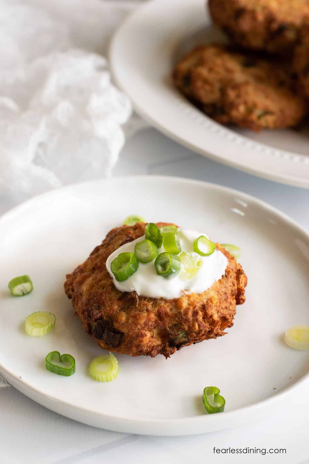 A salmon patty on a plate. It is topped with sour cream and fresh sliced scallions.