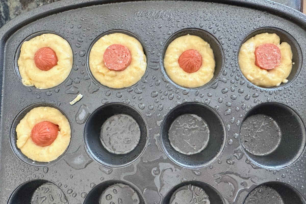A mini muffin pan sprayed with oil. A few cups have muffin batter and hot dog.
