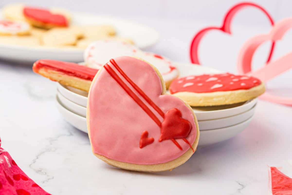 A decorated heart cookie on its side next to plates of cookies.