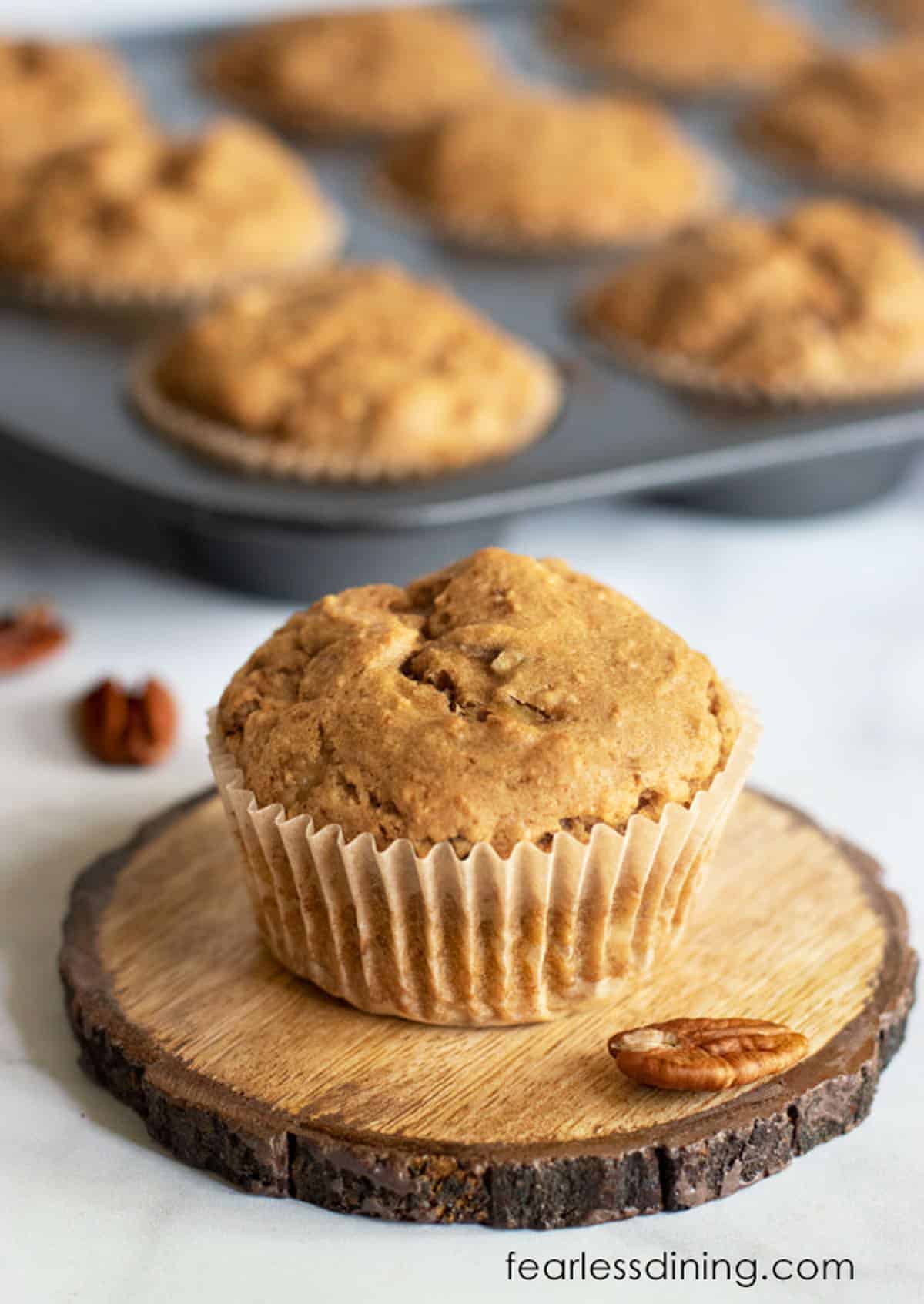 A gluten free banana muffin on a wooden coaster next to the pan of muffins.