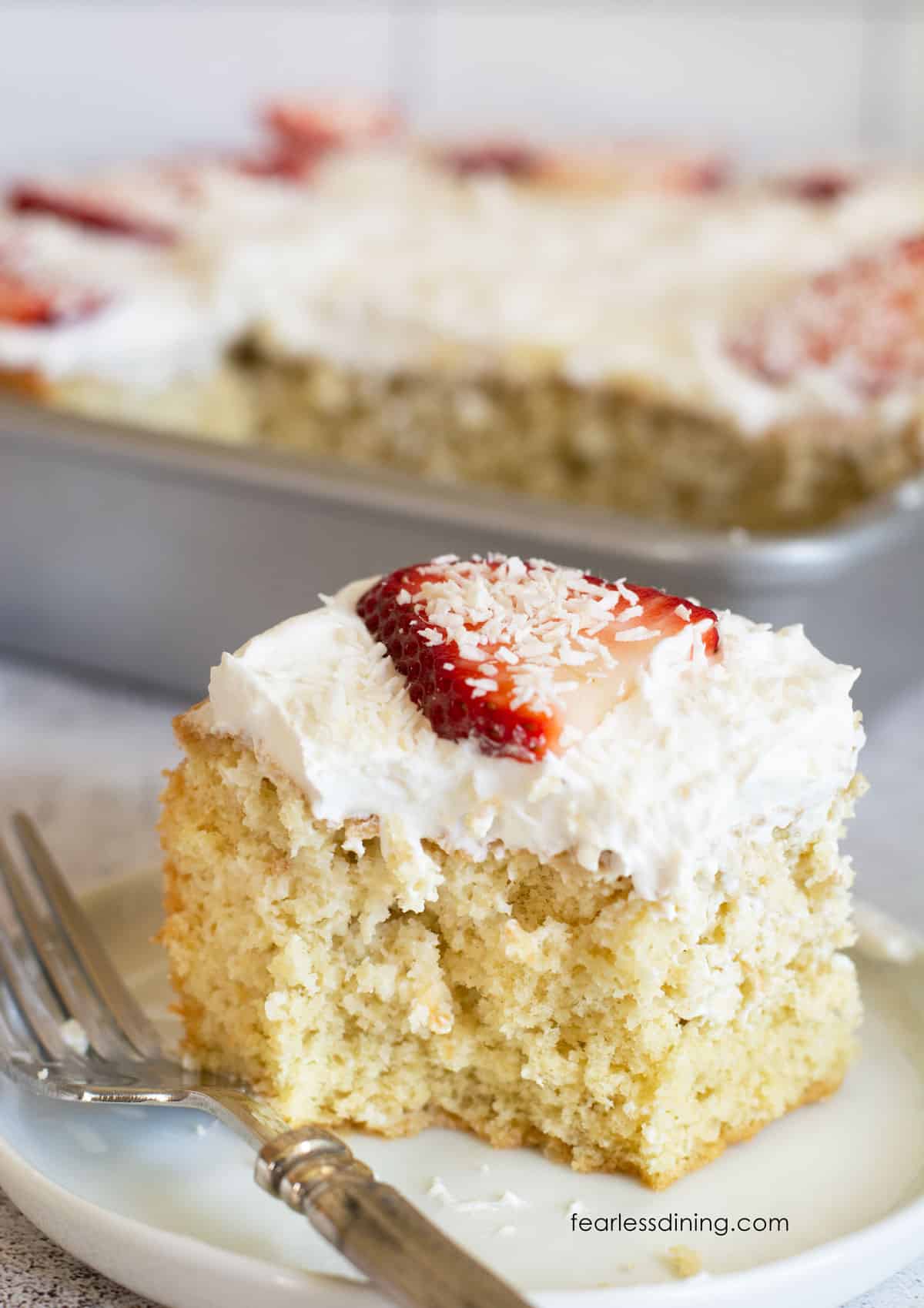 A slice of gluten free tres leches on a plate. It is topped with fresh whipped cream, coconut, and a strawberry.