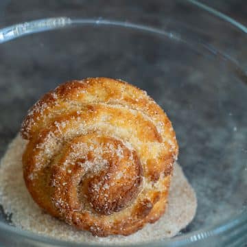A baked cruffin in a bowl of cinnamon sugar.