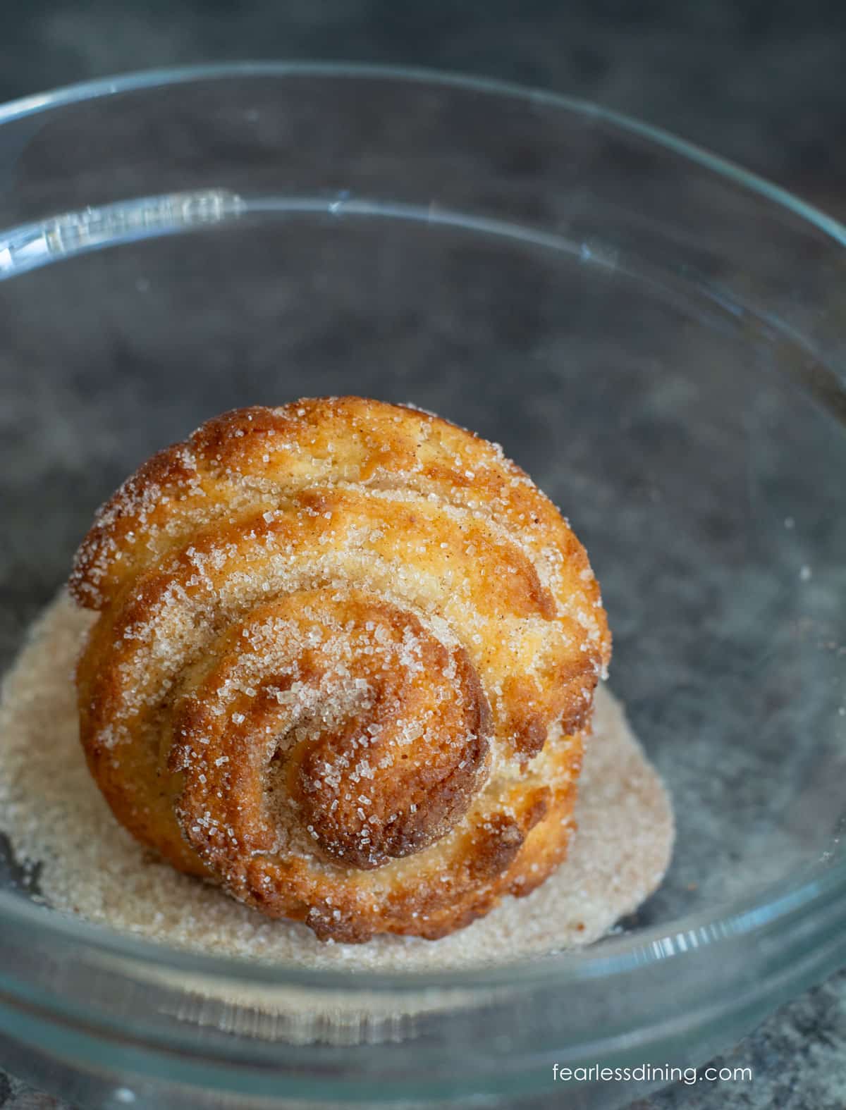 A baked cruffin in a bowl of cinnamon sugar.