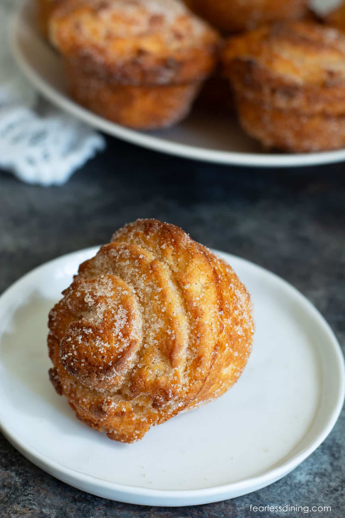 A gluten free cruffin on a small white plate next to a platter of cruffins.