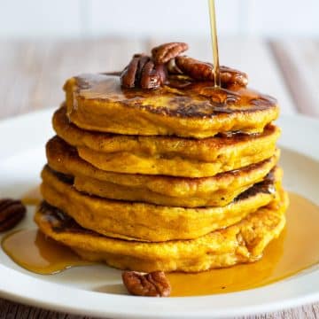 A stack of five gluten free pumpkin pancakes on a white plate. Maple syrup is being drizzled over the top.