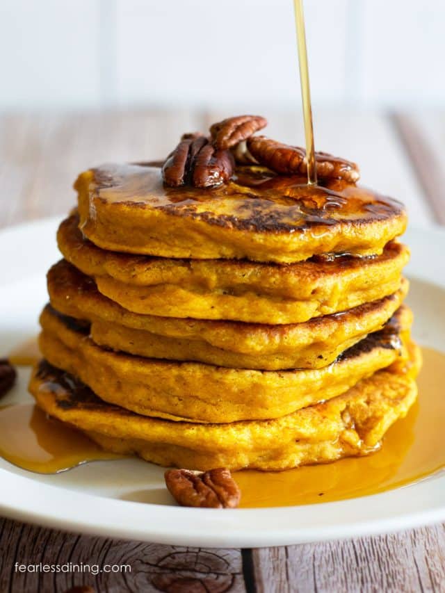 A stack of five gluten free pumpkin pancakes on a white plate. Maple syrup is being drizzled over the top.