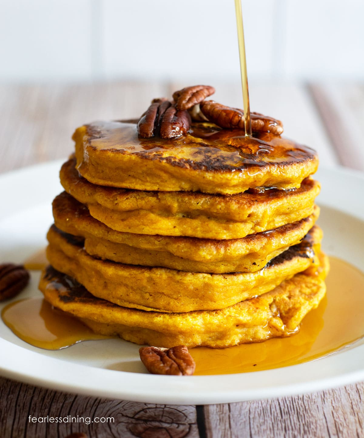 A stack of five gluten free pumpkin pancakes on a white plate. Maple syrup is being drizzled over the top.