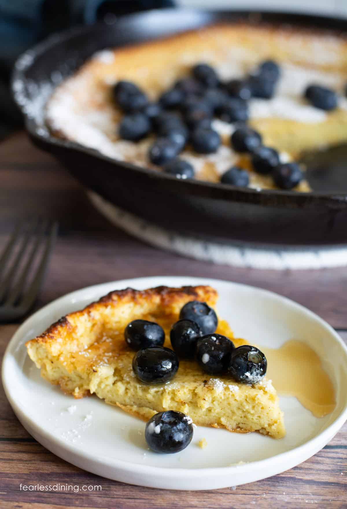 A slice of puff pancake on a plate. It is topped with blueberries and maple syrup.