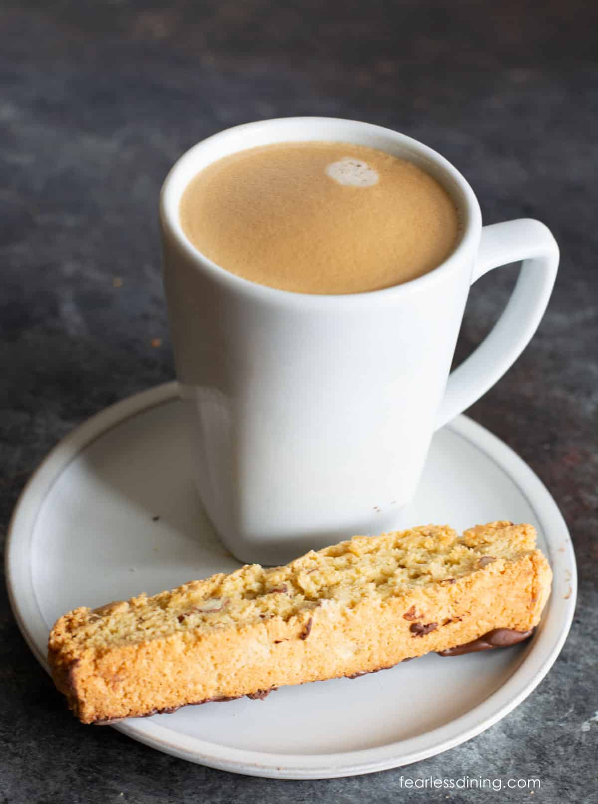 A slice of almond biscotti on a small plate with a tall cup of coffee.