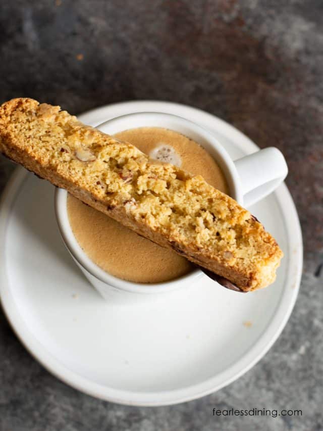 A slice of gluten free almond biscotti sitting on the rim of a coffee cup.
