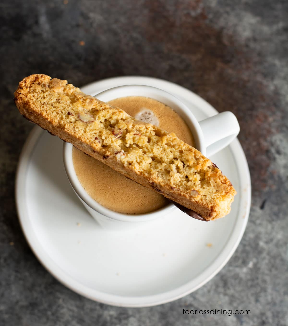 A slice of gluten free almond biscotti sitting on the rim of a coffee cup.