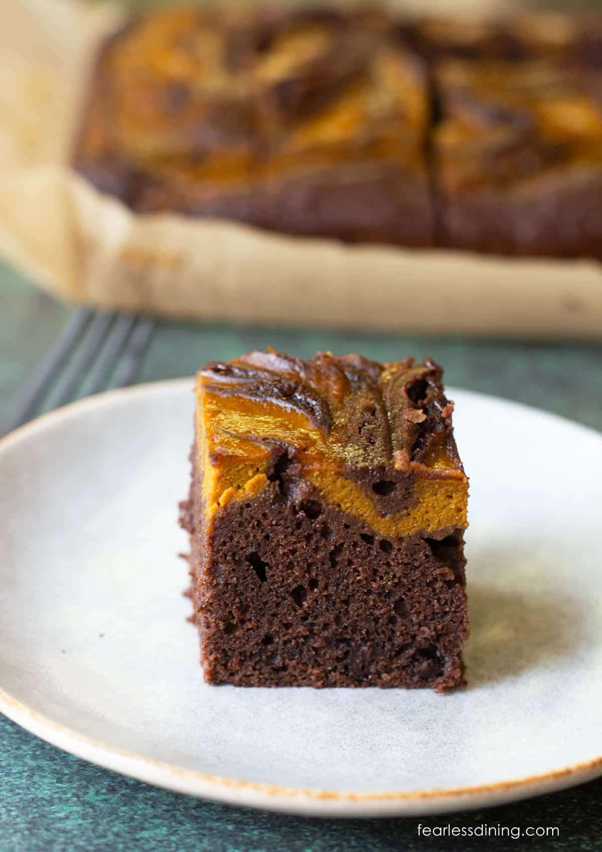 A gluten free pumpkin swirl brownie on a plate in front of the tray of brownies.